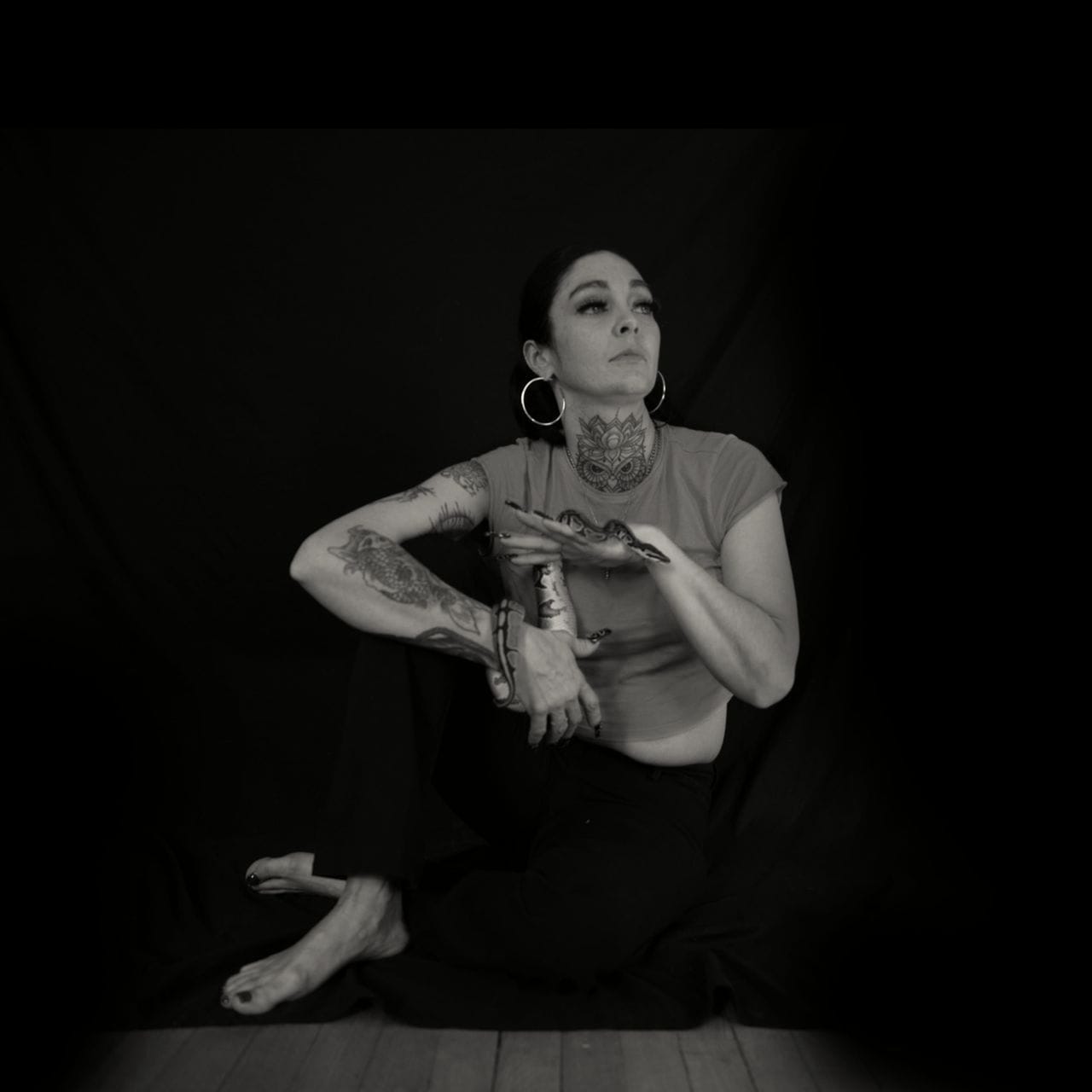 Andrea Martin sits on the floor in front of a dark background with her arm propped up on her right knee. She’s also holding her pet snake, which is wrapped around her right wrist. A younger woman, she wears a red t-shirt and large hoop earrings, and has her dark hair pulled back.