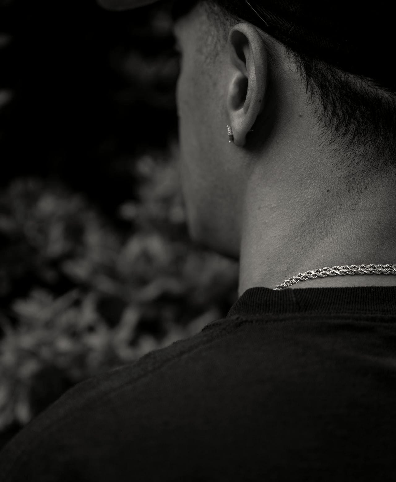 A view of John Doe #141 from the left side of his back, from the chest up to the side of his face. He is wearing a black t-shirt, gold chain around his neck, and a diamond earring in his left ear, and a black baseball cap. His dark hair is cut short.